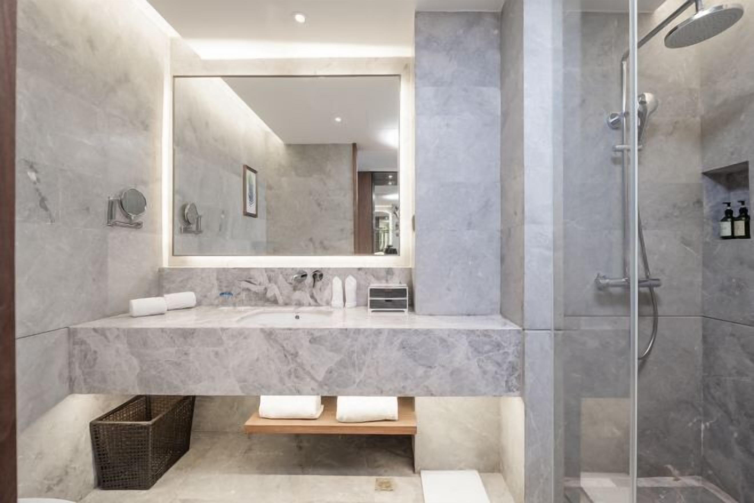 Un baño moderno con tocador de mármol, un espejo grande, cabina de ducha y un pequeño estante de madera.