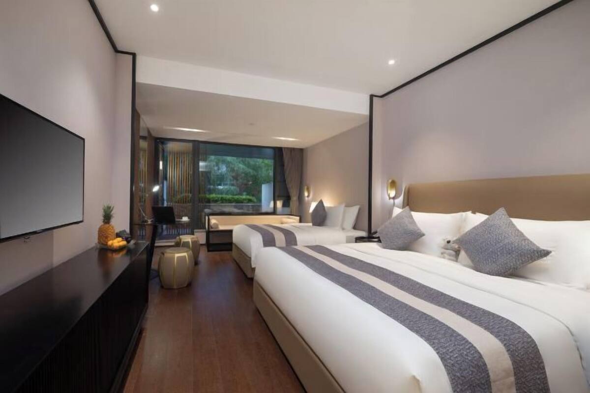 A modern hotel room with a large bed, a TV, a desk with a chair, and a view of greenery outside.