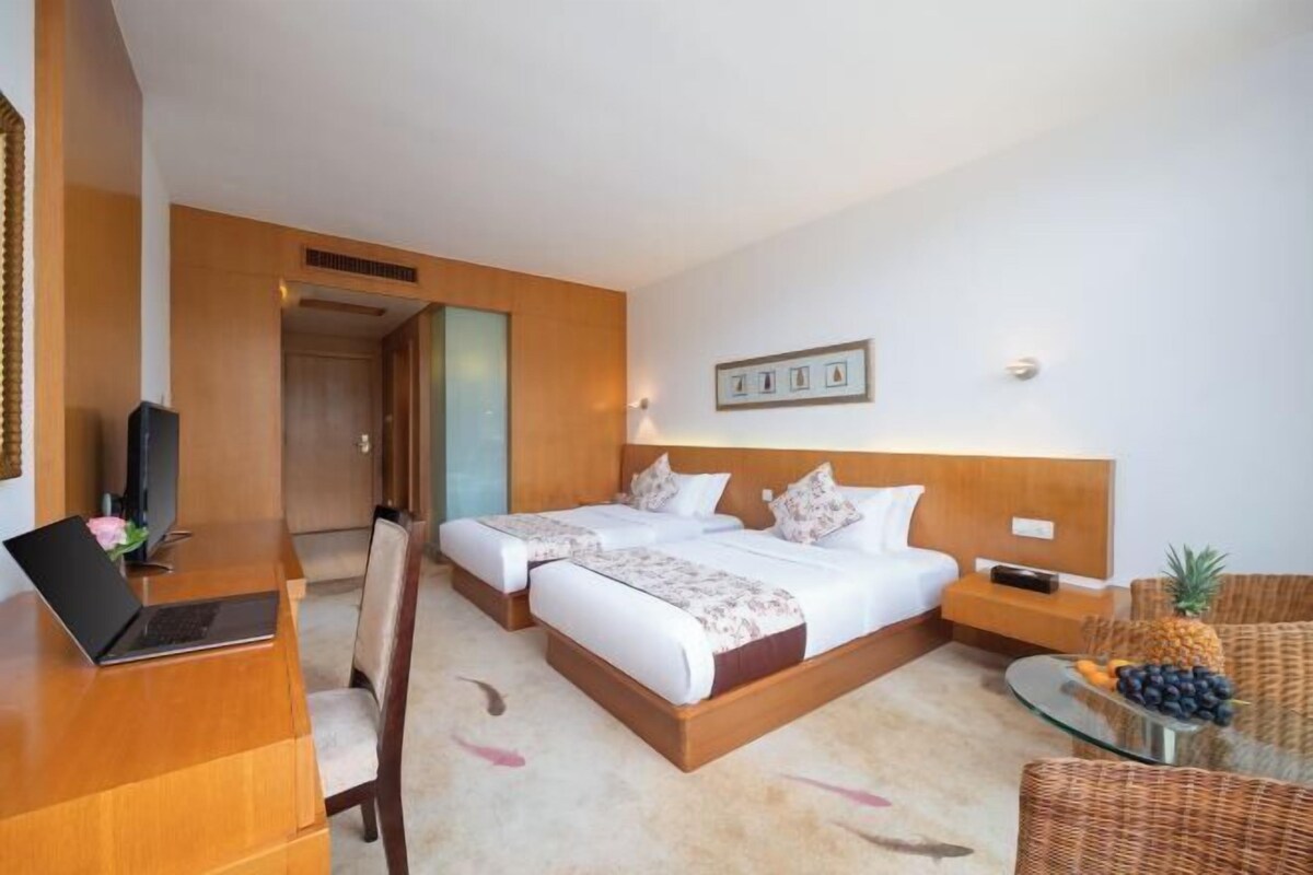 A hotel room with two beds, a desk with a laptop, a TV, and a wicker chair with fruit.
