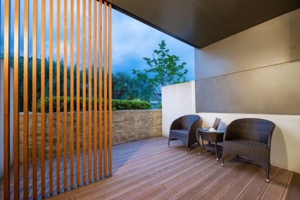An outdoor patio with wooden flooring, two wicker chairs, a small table, and a wooden partition.