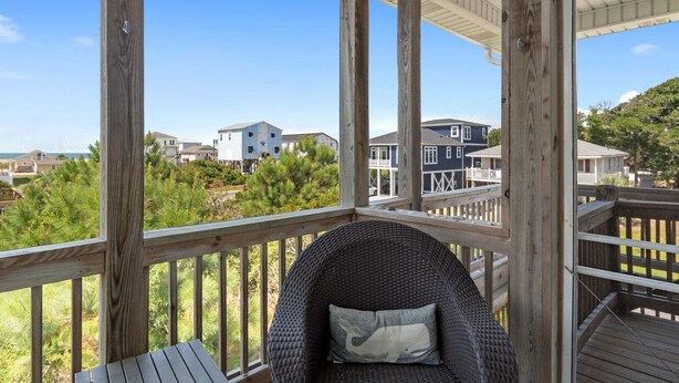 Una terraza de madera con una silla de mimbre y una mesa, con vistas a una zona residencial con casas y vegetación.