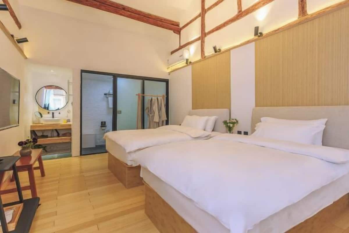 A hotel room with two beds, wooden flooring, a wooden headboard, a bedside table, a mirror, and a bathroom visible through an open door.