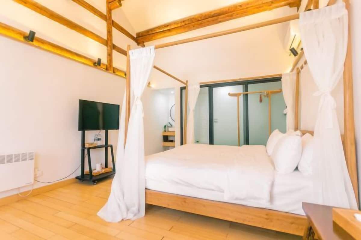 A bedroom with a wooden bed frame, white bedding, and a canopy. There is a TV on a stand to the left and a window with curtains to the right.