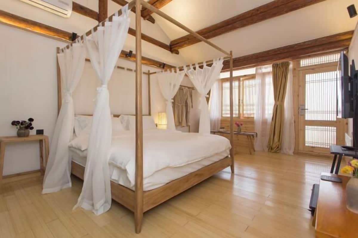 A four-poster bed with sheer curtains, a wooden floor, a bedside table with a lamp, and a window with curtains.