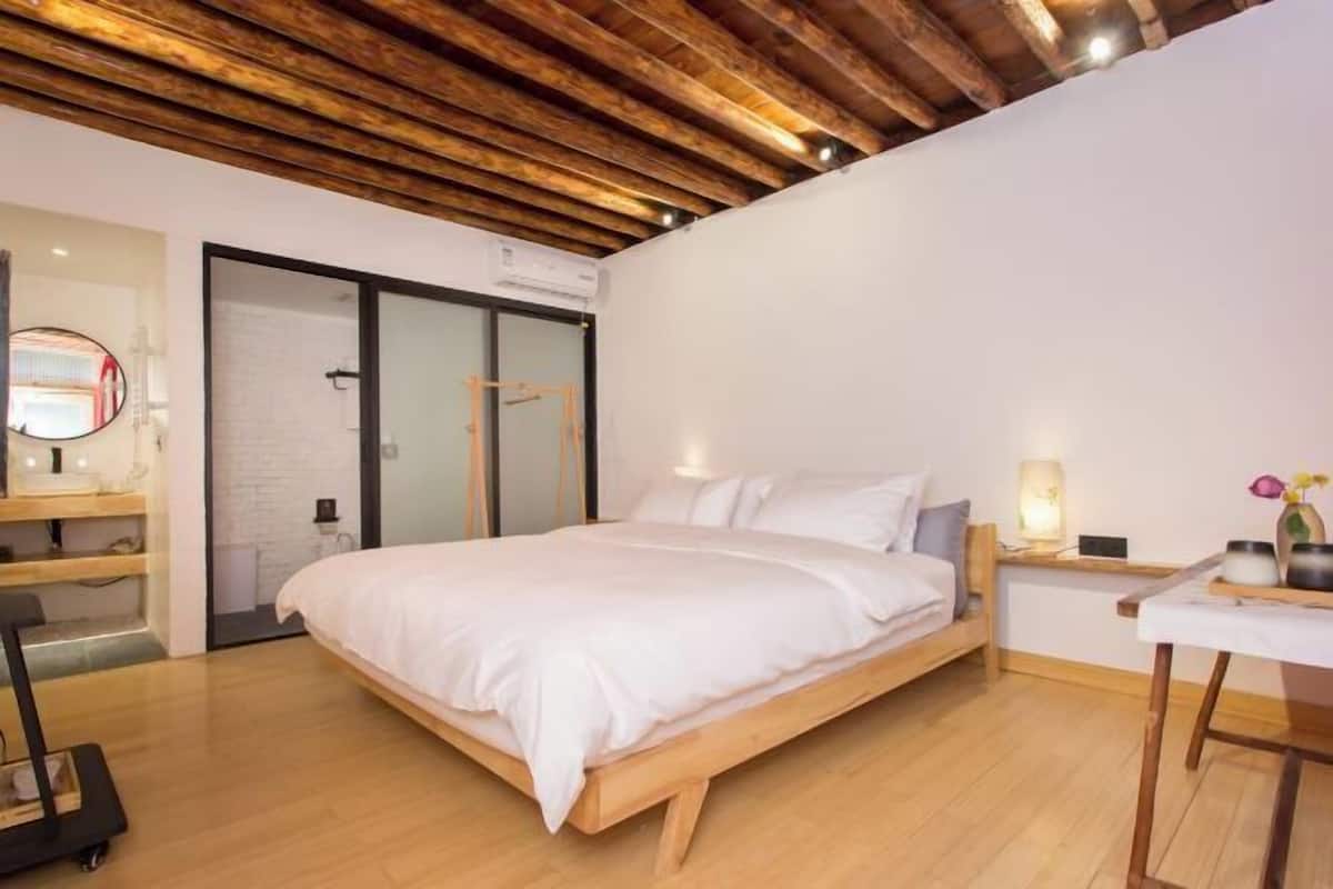A modern bedroom with a wooden ceiling, a bed with white bedding, a desk with a lamp, and a small table with flowers.