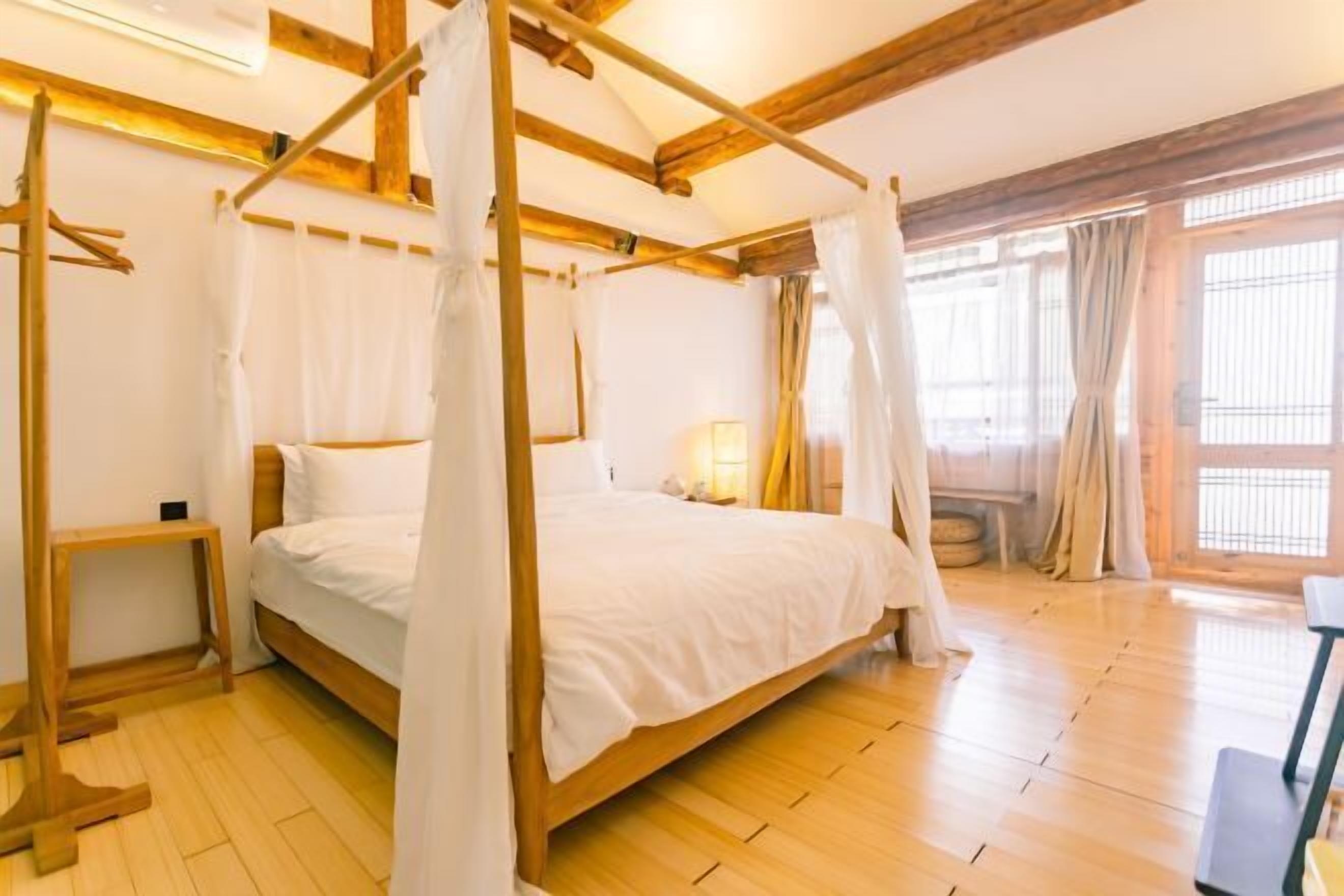 A room with a wooden four-poster bed, a bedside table, and a window with curtains.