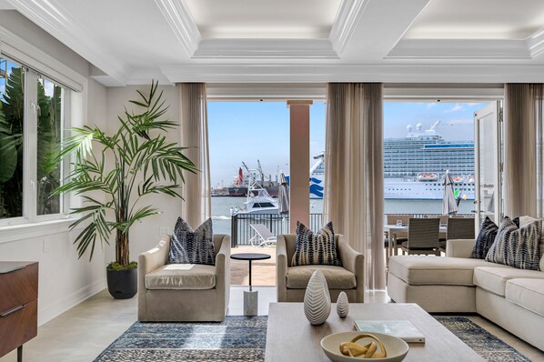 A spacious living room with a large potted plant, two beige sofas with patterned cushions, a coffee table with decorative items, and a view of a cruise ship and marina through large windows. - Million Dollar Views / Private beach / Heated pool / Walk to Beach / Harbourview Key | VlLLAS (Fort Lauderdale)