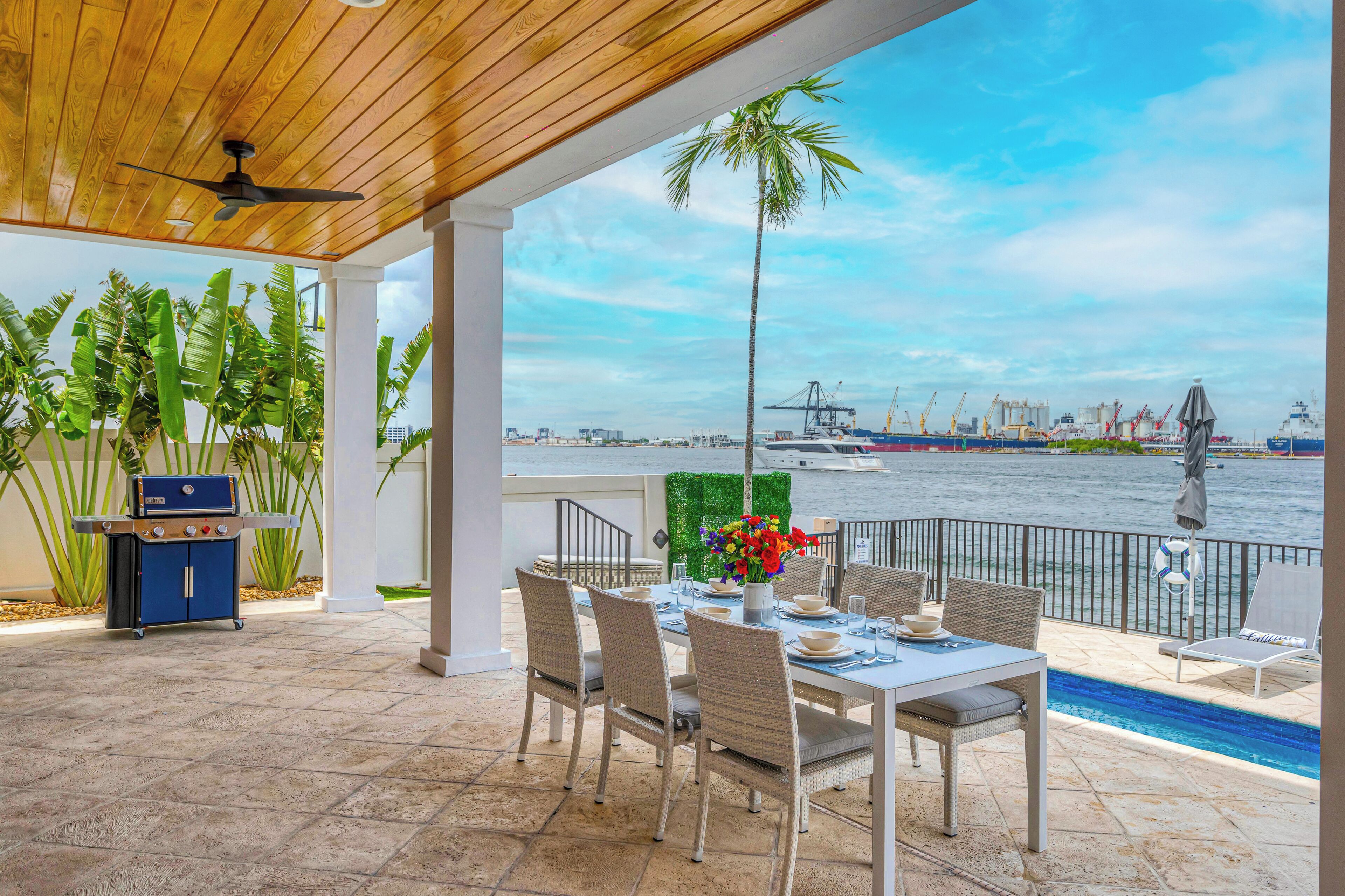 An outdoor patio with a dining table, chairs, a grill, and a view of the water and port.