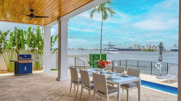 An outdoor patio with a dining table, chairs, a grill, and a view of the water and port.