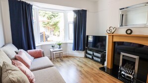 A living room with a sofa, TV, fireplace, and a window with curtains. - 13 The Crescent (Colwyn Bay)