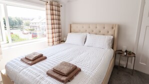A neatly made bed with two folded towels, a window with a view of a street and buildings, and a small bedside table with a lamp and a book. - 13 The Crescent (Colwyn Bay)