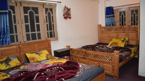 A room with two wooden beds, one with a patterned bedspread and the other with a floral one. There is a small wooden nightstand between the beds. - Hotel 1947 (Gilgit)