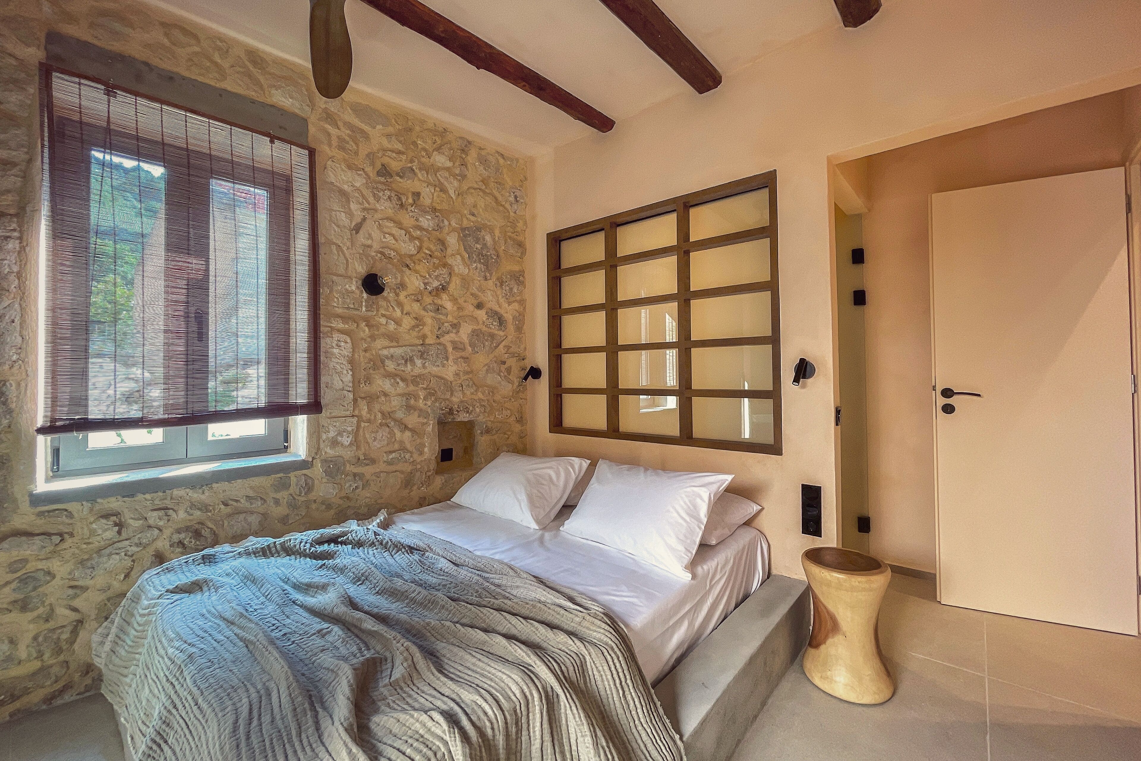 Une chambre avec un mur en pierre, une grande fenêtre avec des stores, un lit avec du linge de maison blanc, une tête de lit en bois et une porte dotée d’une poignée en bois.