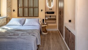 A bedroom with a large bed, a wooden ceiling, a stone wall, a wooden floor, a built-in bench, and a small bathroom with a mirror and sink. - Kaliva Residence (Apokoronas)