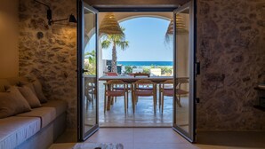A room with a stone wall, a sofa, a dining table with chairs, and a view of the sea through an open door. - Kaliva Residence (Apokoronas)