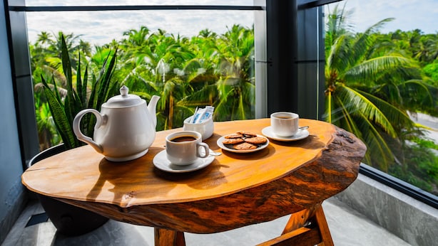 A wooden table with a teapot, cups, and cookies, set against a backdrop of lush green palm trees through large windows. - Maa Thundi (Fuvahmulah)