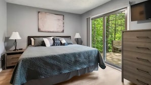 Un dormitorio con cama, dos mesitas de noche con lámparas, un ventanal con vistas a la vegetación y un televisor montado en la pared.