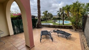 A covered patio with two sun loungers, a palm tree, and a view of a swimming pool and greenery.