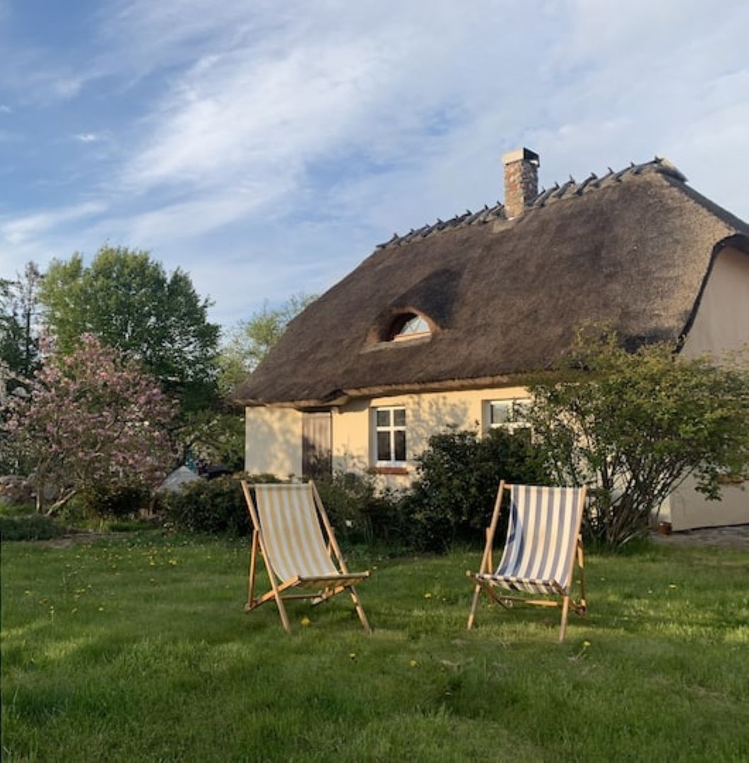 Een huis met rieten dak, twee houten stoelen in de voortuin en bloeiende bomen.