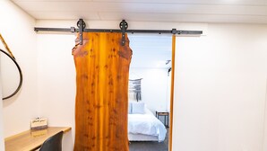 Ein Zimmer mit Holztür, Schreibtisch mit Stuhl, einem Bett und einem kleinen Tisch.