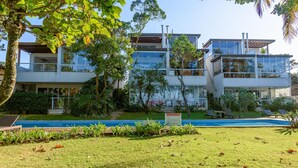 A modern multi-story building with a pool, surrounded by lush greenery and palm trees. - Pé na areia com charme e elegância (São Sebastião)