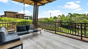 Ein Balkon im Freien mit Holzdeck, schwarzen Sitzmöbeln mit Polstern und Blick auf einen angelegten Garten sowie Gebäude im Hintergrund.