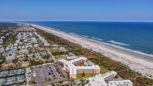 An aerial view of a coastal area with a beach, a parking lot, and a hotel with multiple floors and a swimming pool.