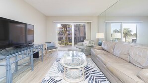 A living room with a TV, a glass coffee table, a sofa, and a chair. There is a balcony with a view of palm trees and buildings.