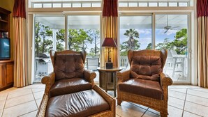 A room with two leather armchairs, a small table with a lamp, and a large sliding glass door leading to an outdoor area with trees and a view of a house. - Let Camelot become your escape from realty. (St. George Island)