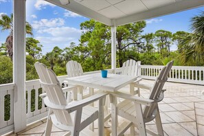 A covered outdoor patio with a white table and chairs, surrounded by a white fence, overlooking a lush green landscape with trees. - Let Camelot become your escape from realty. (St. George Island)