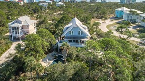 A coastal residential area with multiple houses, a large house with a wrap-around porch, and a view of the ocean. - Let Camelot become your escape from realty. (St. George Island)