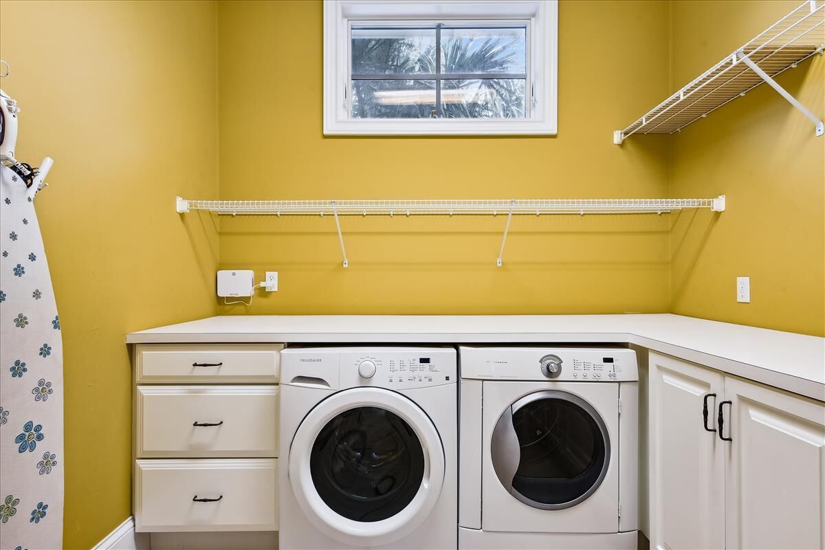 A laundry room with two washing machines, a drying rack, and a window with a view of palm trees.