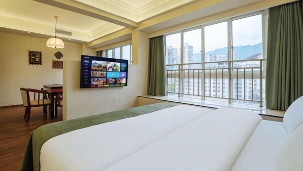 Een hotelkamer met een groot bed, een aan de muur bevestigde televisie, een bureau en uitzicht op een stadsgezicht door grote ramen.