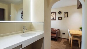 Een moderne badkamer met een grote spiegel, een wastafel en een badkamermeubel. In de aangrenzende kamer staat een bruine leren bank en een houten eettafel.