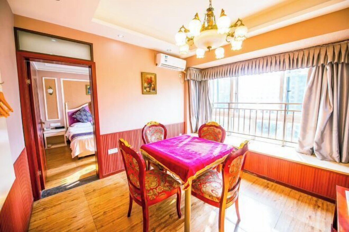 Um quarto com piso de madeira, uma mesa de jantar com cadeiras, um lustre, uma janela com cortinas e uma porta que dá para outro cômodo.