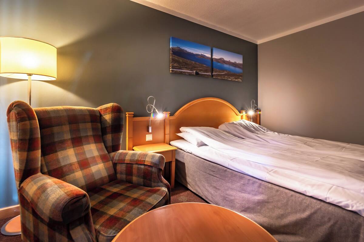 A hotel room with a bed, a chair, a nightstand, a lamp, and a wall-mounted picture.