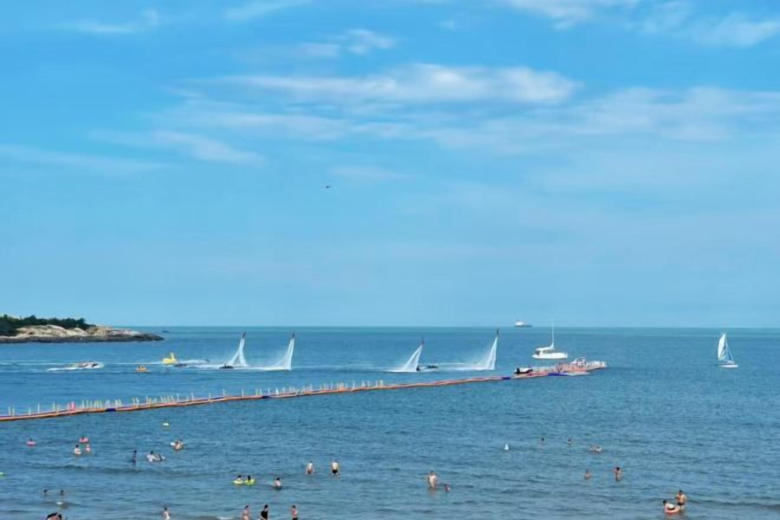 Escena costera con un muelle que se adentra en el agua, barcos y gente nadando.