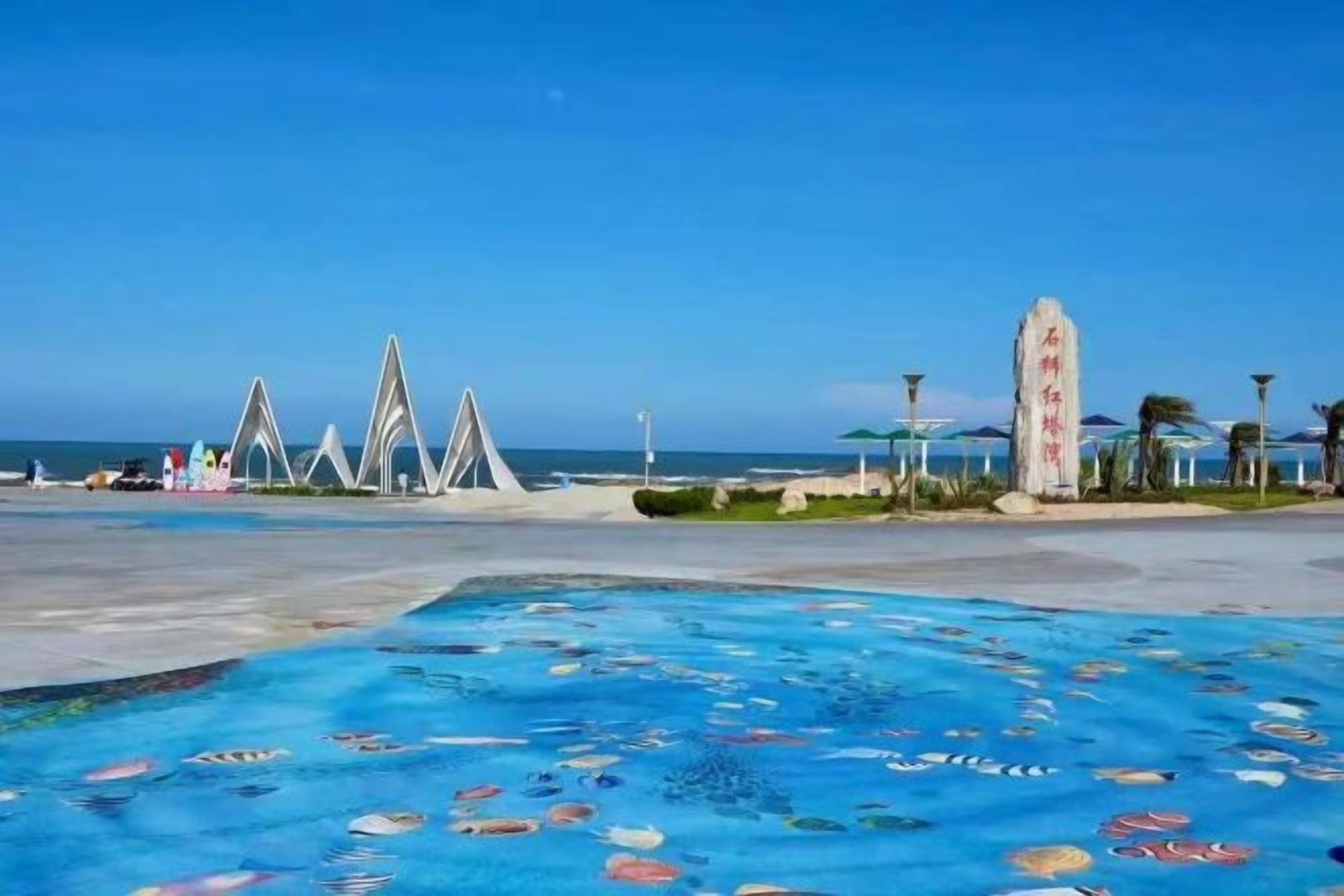 Una playa con una piscina estampada, cielo despejado y una serie de estructuras blancas al fondo.