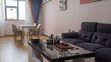 Ein modernes Wohnzimmer mit einem dunklen Sofa, einem Essbereich mit Tisch und Stühlen sowie einer Küchenzeile mit Kaffeemaschine und Snacks.
