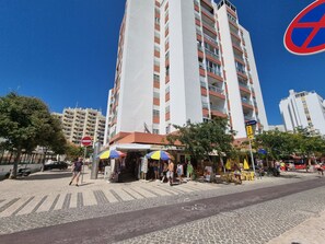 Un edificio de varios pisos con balcones y una zona peatonal con tiendas y mesas al aire libre.