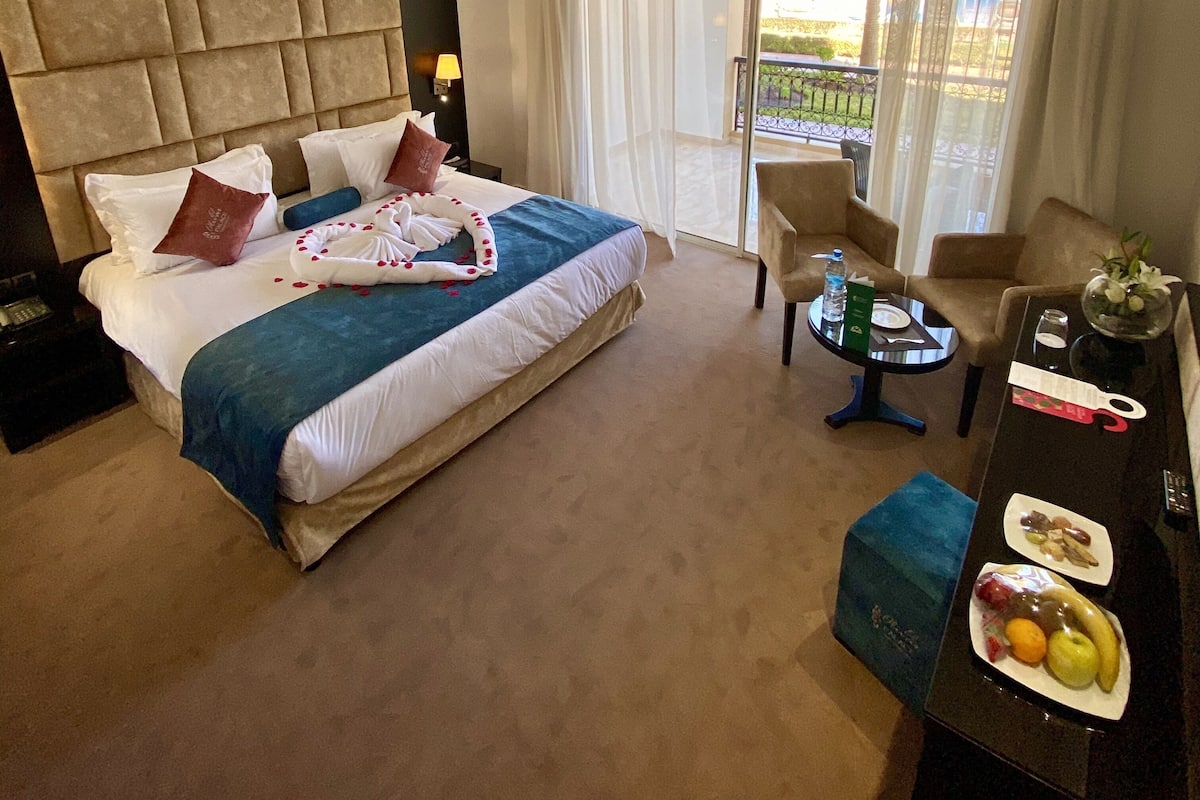 A hotel room with a large bed, a desk with a chair, a small table with a bottle and a plate of fruit, and a view of the outdoors through the window.
