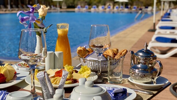 Un bufé de desayuno con diversas opciones de comida y bebidas, una piscina al fondo y sillones de descanso a orilla de la piscina.