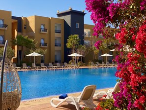 Un área junto a la piscina con sillones de descanso y sombrillas, una piscina azul cristalina y un edificio con balcones y una torre.