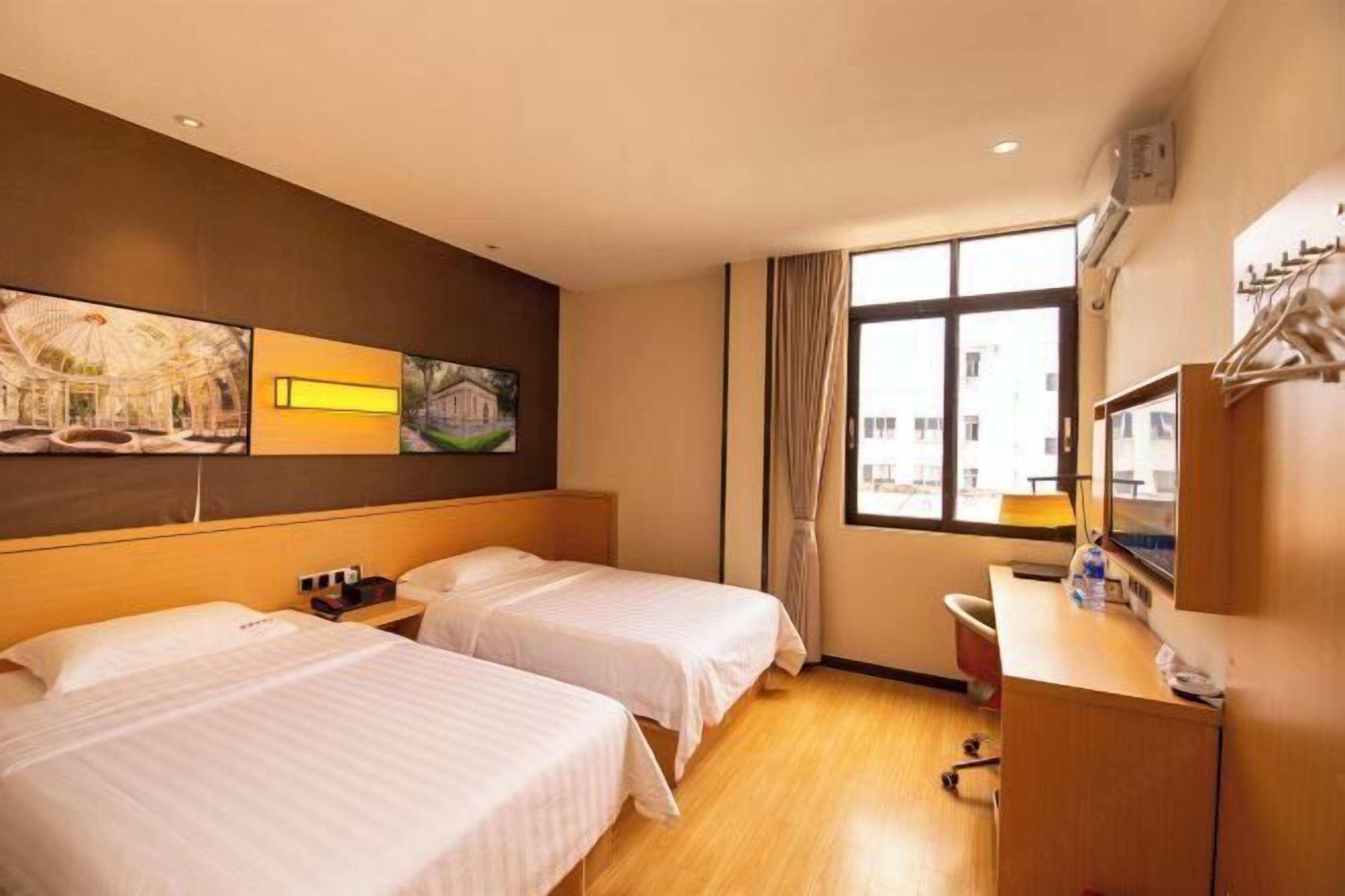 A hotel room with two beds, a desk, a chair, a window with a view, and artwork on the wall.