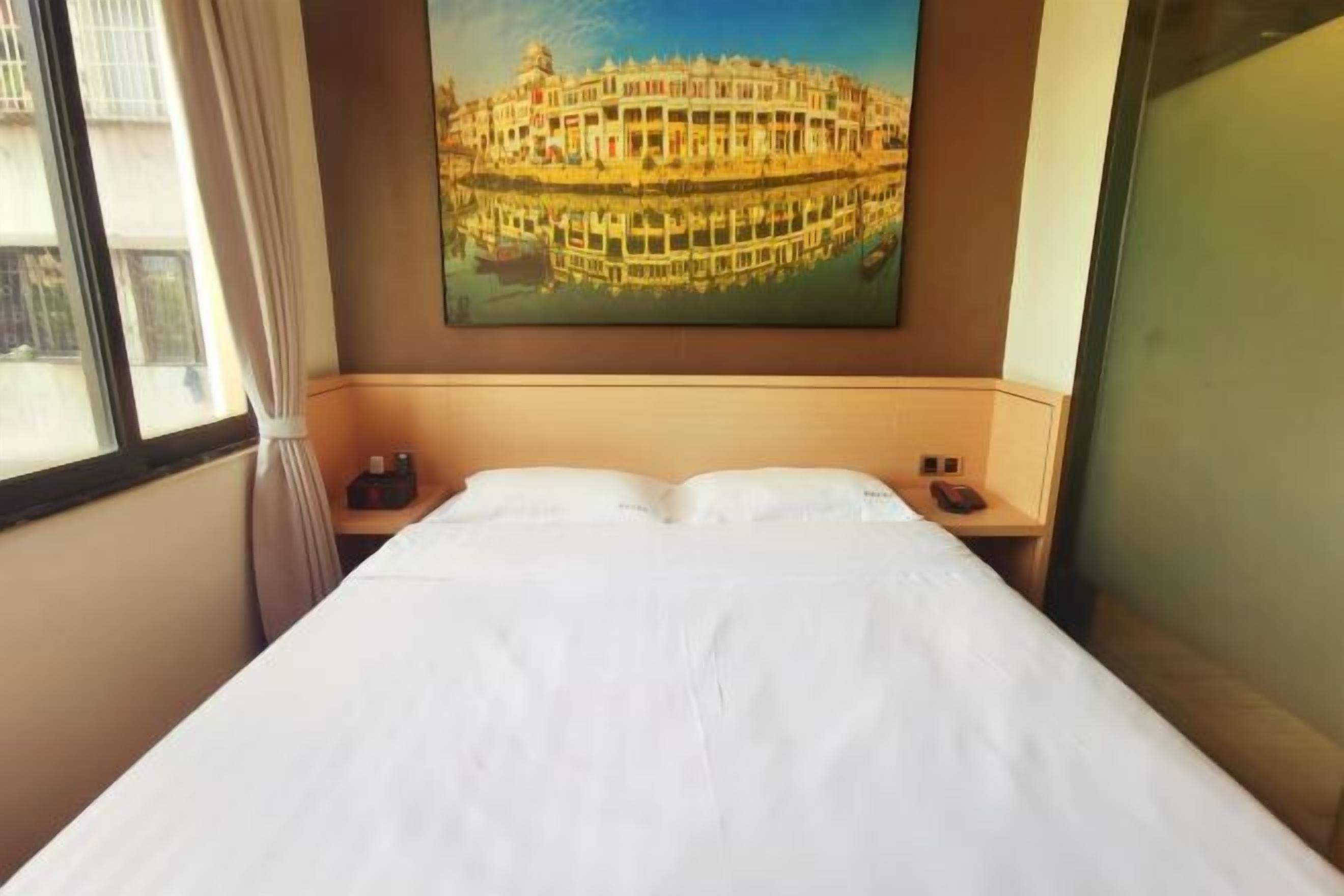 A hotel room with a bed, a nightstand, a painting of a building, and a window with curtains.