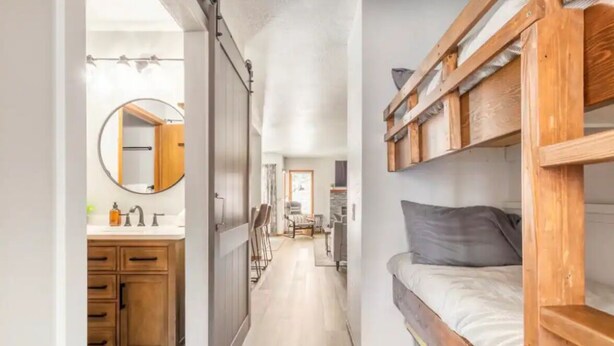 Um corredor estreito que dá para um quarto com uma cama beliche, um armário de madeira e um espelho redondo no quarto ao lado.