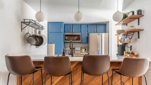 Uma cozinha moderna com armários azuis, uma ilha central com banquetas, panelas penduradas e piso de madeira.