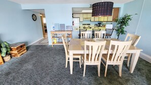 Un coin repas avec une table et des chaises en bois, une cuisine à l’arrière-plan et une plante sur la droite.