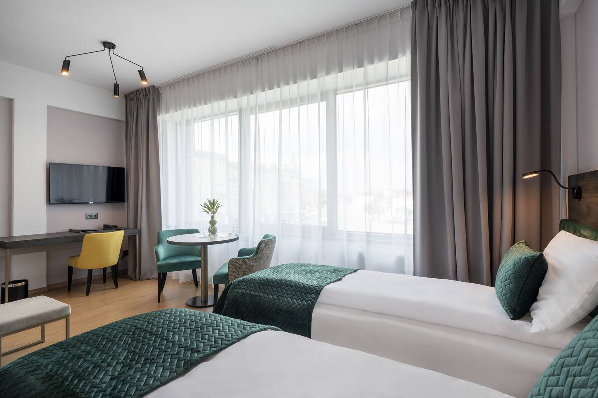 Hotellihuone, jossa on kaksi sänkyä, työpöytä, tuoli, televisio ja ikkuna, jossa on verhot.