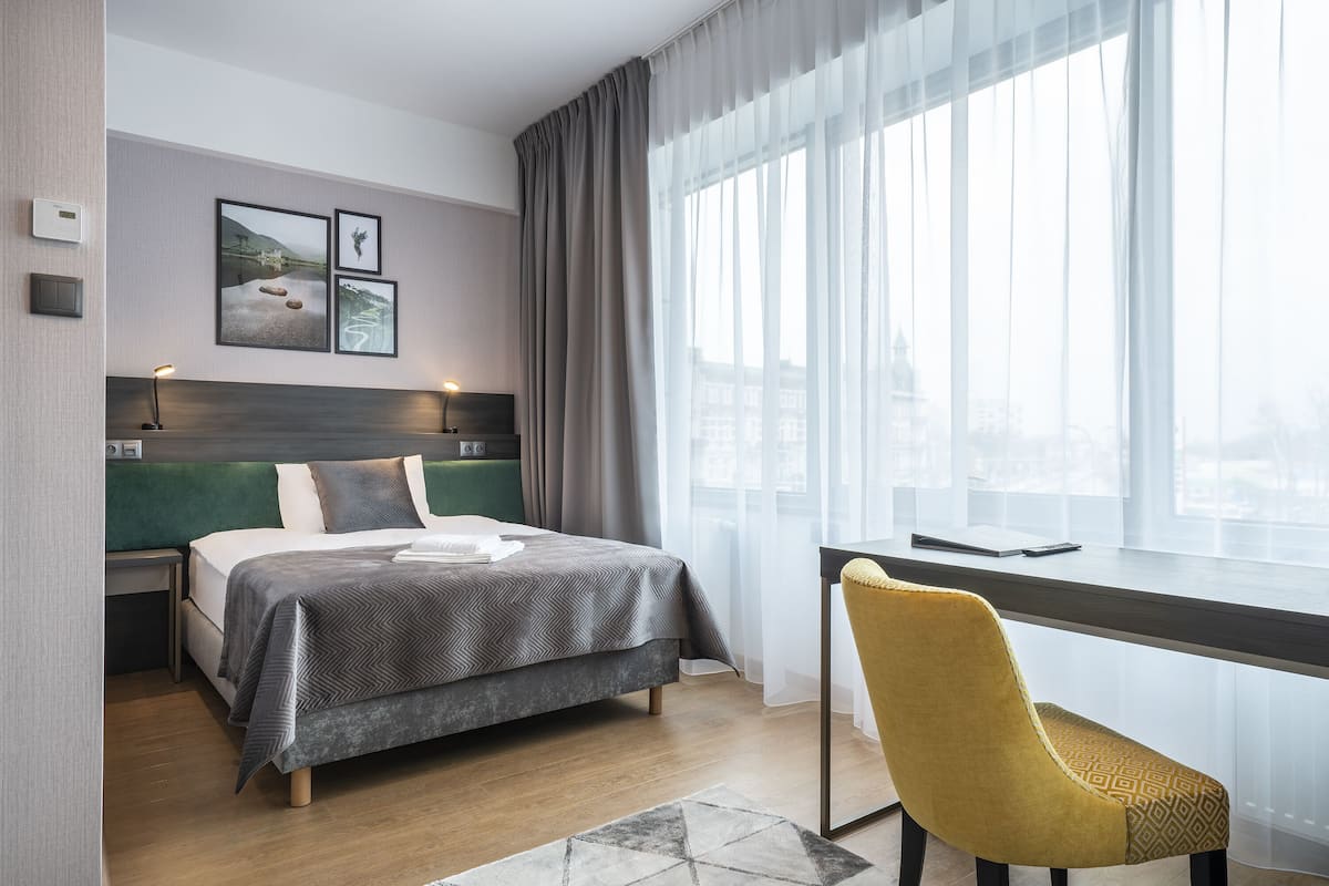 Moderni hotellihuone, jossa on sänky, työpöytä ja tuoli. Seinällä on kolme kehystettyä kuvaa, ikkunassa on verhot ja ikkunasta avautuu näkymä kaupunkiin.
